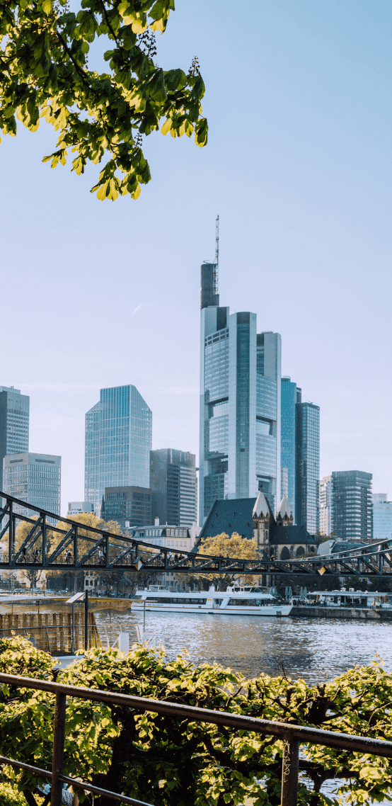 Frankfurt skyline with modern skyscrapers, a bridge over the Main River, and green foliage.