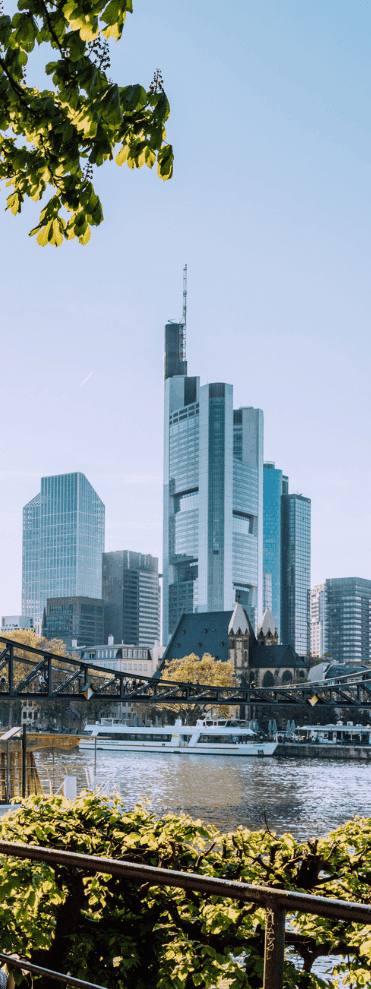 Modern skyscrapers of Frankfurt city skyline beside a river, framed by green foliage.